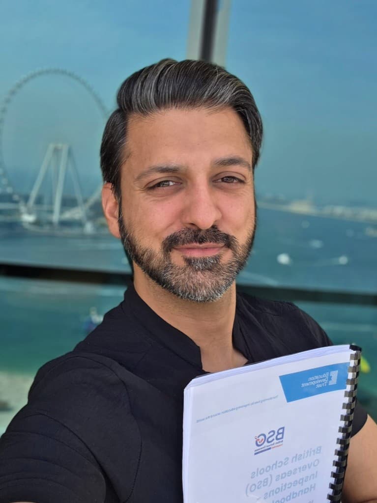 Adnan, smiling, holding a British Schools Overseas inspection handbook, in a black collared shirt with a bright window and water behind him.