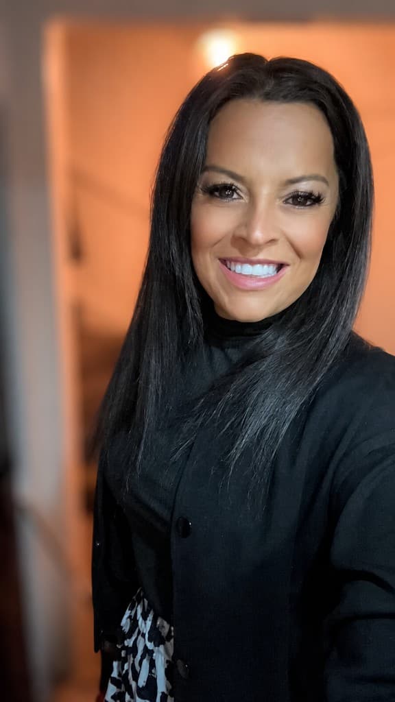 Danielle, smiling, wearing a black top and jacket in a warmly lit hallway.