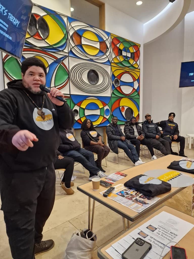 A Teacher's Surgery speaker at the One For All event, with a seated panel, colourful community artwork, and event materials on the table.